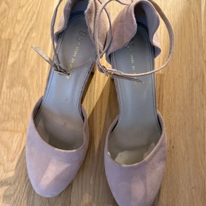 Ljusrosa mocka pumps med klack - Snygga ljusrosa pumps i mjuk mocka från DS Shoes by Caprice. Skorna har rund tå, hög klack och justerbara remmar med guldfärgade spännen runt ankeln. Perfekta för dig som vill ha en chic och feminin look med extra höjd.