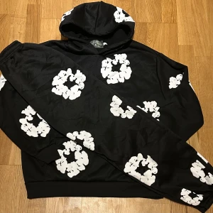 Denim tears hoodie och byxor  - Svart hoodie från denim tears med stora vita blommor tryckta över hela tröjan. Tröjan har huva, magficka och långa ärmar. Mönstret är riktigt unikt och ger en cool vibe. Perfekt för dig som vill sticka ut med en streetstyle-look.