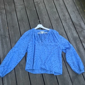 Blå blus med broderade hålmönster - Säljer en ljusblå blus med v-ringning och långa ärmar. Blusen har broderade hålmönster i blommiga former över hela tyget och är gjord i mjuk bomull. Perfekt för dig som gillar detaljer och vill ha en chill men snygg look.