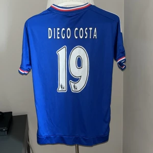 Chelsea Diego Costa #19 blå matchtröja - Säljer en officiell Chelsea matchtröja med Diego Costa och nummer 19 på ryggen. Tröjan är blå med vita och röda detaljer, har smala svarta ränder och korta ärmar. Märkt med Yokohama Tyres på bröstet och Chelsea FC-emblem. Tillverkad i polyester för skön känsla.