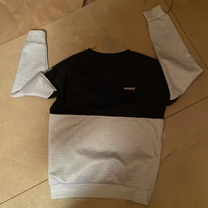 Svart och grå sweatshirt från McKenzie - Säljer en stilren sweatshirt från McKenzie i storlek L för 12-13 år. Tröjan har en cool färgblock-design med svart upptill och ljusgrått nedtill samt på ärmarna. Rund hals och mjukt material som är skönt mot huden. Perfekt för chill dagar eller häng med kompisar.