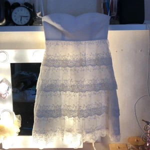 Vit axelbandslös spetsklänning - Superfin vit axelbandslös klänning med flera lager spets och blommigt mönster. Kjolen är kort och har volangdetaljer som ger en romantisk vibe. Perfekt för student eller sommarens alla tillfällen. Sitter snyggt med en slim passform och har en feminin look.