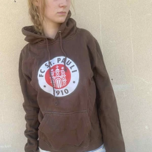 Brun FC St. Pauli hoodie med tryck - Säljer en brun hoodie från FC St. Pauli med stort vitt och rött klubbmärke på bröstet. Tröjan har huva med snörning, magficka och långa ärmar. Listad som XL men sitter mindre, personen på bilden är 167cm.