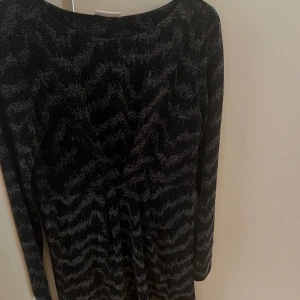 Svart glittrig långärmad klänning - Snygg svart klänning med glittrigt zebramönster och lång ärm. Klänningen har en v-ringad front och är tillverkad i ett glittrigt syntetmaterial som ger en festlig vibe. Perfekt för dig som vill sticka ut med coola detaljer och mönster.