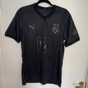 Svart dortmund fotbollströja - Snygg svart Borussia Dortmund fotbollströja från Puma med diskret mönster och silvriga detaljer. Kortärmad med V-ringad krage, BVB-logga och 1&1 sponsortryck på bröstet. Tillverkad i lätt och ventilerande polyester, perfekt för match eller träning.