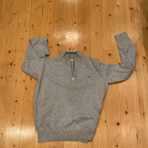 Grå half zip tröja från GANT - Säljer en stilren grå tröja från GANT med half zip och ribbad krage med svarta ränder. Tröjan har lång ärm och är tillverkad i mjuk bomull, perfekt för chill dagar. Diskret GANT-logo på bröstet och klassisk passform. Priset kan diskuteras