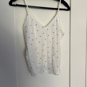 Vit linnetopp med röda hjärtan - Supersöt vit linnetopp med tunna axelband och små röda hjärtan över hela tyget. Toppen har spetskant vid halsen och en liten rosett framtill. Materialet är mjukt och ribbat, funkar även perfekt som ett nattlinne/pyjamas linne. Aldrig använd