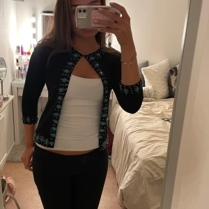 Svart kofta med blåa stenar - Svart väst med trekvartsärm och blå stenar med guld detaljer längs kanterna och ärmsluten. Snygg och unik design som sticker ut, perfekt för att lyfta en enkel outfit.