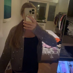Grå kofta med pärlor, croppad - Superfin grå kofta med pärldetaljer över hela tröjan. Modellen är croppad och har långärmade, lite loose ärmar. Stickad i mjukt material som känns skönt mot huden. Perfekt att slänga över en topp för en chill och snygg look. passar xs🩷