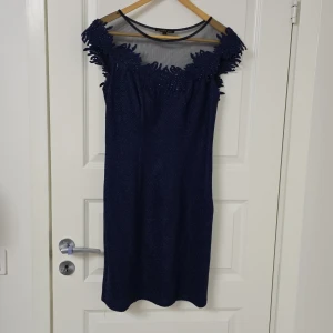 Marinblå kortklänning med spets - Snygg marinblå kortklänning från Aurelia med transparent mesh upptill och dekorativ spets med pärlor runt halsringningen. Klänningen har en figurnära passform och är ärmlös, perfekt för dig som vill sticka ut med elegans och coola detaljer.