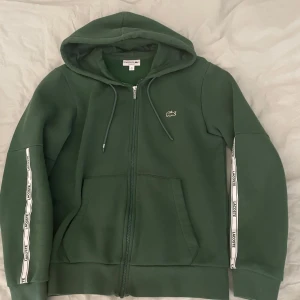 Lacoste hoodie  -  Grön hoodie från Lacoste med dragkedja och huva. Jackan har vita band med Lacoste-logga längs ärmarna, snörning vid huvan och två stora fickor framtill.