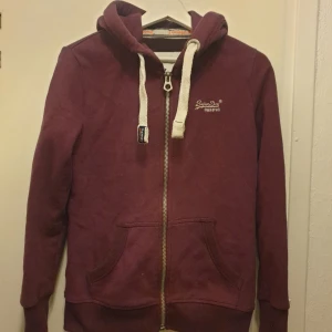 Vinröd hoodie från Superdry - Säljer en vinröd hoodie från Superdry med vit snörning och dragkedja framtill. Tröjan har huva, två fickor och broderad logga på bröstet. Perfekt för chill dagar och snygg till jeans. Mjuk bomullskvalitet och långärmad modell. Färgen gör sig inte rätta på bilden. Använd enstaka gånger. Storlek S. 