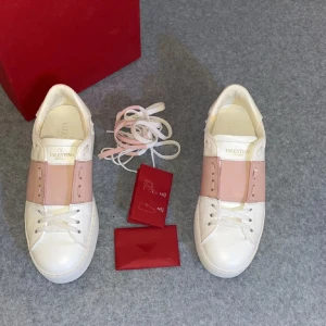 Valentino Garavani Rockstud sneakers - Säljer ett par Valentino Garavani Rockstud sneakers i vitt och ljusrosa skinn. Skorna har en bred rosa panel över snörningen och ikoniska pyramidnitar på hälen. Platt sula och rund tå ger en clean och modern vibe. Perfekt för dig som gillar lyxiga detaljer och stilren design.