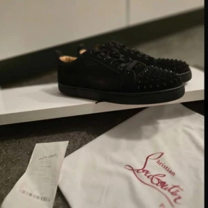 Svarta Christian Louboutin sneakers med nitar - Säljer ett par svarta sneakers från Christian Louboutin med ikonisk röd sula och svarta nitar på tån. Skorna har snörning, rund tå och är tillverkade i mocka med läderdetaljer. Insidan är beige och de har en riktigt edgy vibe som sticker ut.