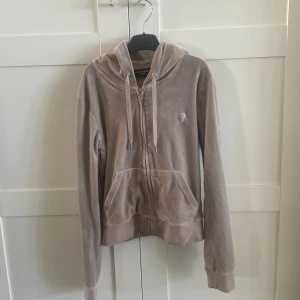 Beige velour hoodie Juicy Couture - Säljer en beige hoodie från Juicy Couture i mjuk velour med huva och snörning. Tröjan har dragkedja med en snygg J-detalj, långa ärmar och fickor framtill. Perfekt för chill dagar och har en klassisk, clean look.