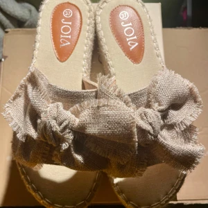 Joia Espandrillos i taupe, strl 37 - Säljer ett par sandaler från Joia i storlek 37. De har en bred plattformssula i flätad beige textil och ovandel i taupe-färgat grovt tyg med stor rosett och fransade kanter. Perfekt för dig som gillar boho-vibe och vill ha något som sticker ut. Bara provad ute ett tillfälle, inget att anmärka på.  Normal i stl. Nypris: 350 sek, Pris nu: 150 sek. Dessa stilrena och lätta espadrillos kombinerar komfort och enkelhet med en modern twist. Tillverkade i 100 % syntetiskt tyg ger de en luftig känsla. 