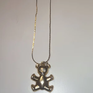 Guldigt halsband med nalleberlock - Snyggt halsband med en gullig nalleberlock i guldton. Kedjan är tunn och berlocken har en lekfull, rundad form som ger ett charmigt intryck. Perfekt för dig som gillar söta detaljer och vill sticka ut lite extra.