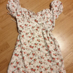 Blommig kortklänning från SHEIN - Supersöt vit kortklänning från SHEIN med puffärmar och blommigt mönster i rött, grönt och lite blått. Klänningen har snörning vid halsen och en feminin passform. Använd 1 gång. Passar M bättre. Man ska nog inte vara över typ 160 om man inte vill ha den allt för kort😅
