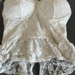 Vit spetskorsett från Bik Bok -  Supersöt vit korsett från Bik Bok i storlek M, täckt med romantisk spets och volangdetaljer nedtill. Axelbandslös bandeau-modell med formade kupor och dragkedja i ryggen. Perfekt för dig som vill ha en feminin och trendig look.