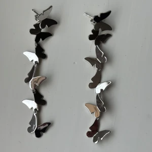 Silvriga örhängen med fjärilar - Säljer ett par coola silvriga örhängen med flera hängande fjärilar i blank metall. Det går att ta av hänget så att man bar har en enkel fjäril. Perfekt för dig som gillar unika detaljer och vill sticka ut. Köpt på butiken Ioaku i Stockholm. Aldrig använda.