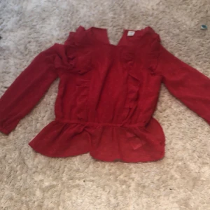 Röd blus med volanger från Lindex - Säljer en röd blus från Lindex i storlek 140. Blusen har lång ärm, volanger längs axlarna och fram, samt prickigt mönster i tyget. Den har en v-ringad öppning bak och ett lätt, luftigt material som känns skönt mot huden. Perfekt för dig som gillar färg och detaljer!