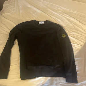 Svart sweatshirt från Stone Island - Svart crewneck sweatshirt från Stone Island med klassisk rund hals och ikonisk logotyp-patch på vänster ärm. Tröjan är tillverkad i mjuk bomull och har ribbade muddar vid ärmslut och nederkant. Perfekt för dig som gillar streetwear och snygga detaljer.