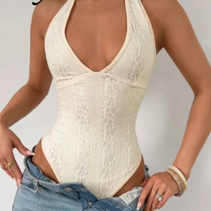 Krämvit spets korsett halterneck - Säljer en krämvit korsett-topp med halterneck och djup v-ringning. Toppen är ärmlös och har öppen rygg, tillverkad i mjuk spets som ger en romantisk vibe. Perfekt att styla med jeans eller kjol för en trendig look. 