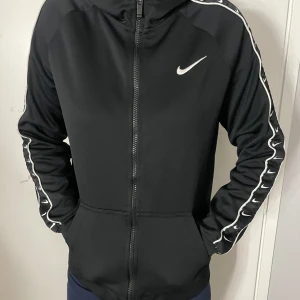 Svart Nike zip-up med huva - Svart zip-up från Nike med huva och hel dragkedja framtill. Snygga vita Nike-loggor längs ärmarna och en vit swoosh på bröstet. Tröjan har fickor och sportig look. Både killar och tjejer kan ha den här.