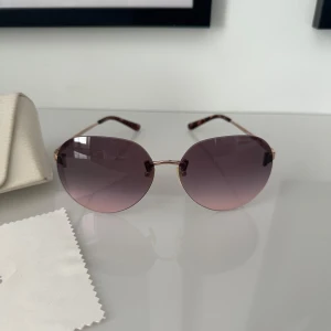 Lila solglasögon från Michael Kors - Snygga solglasögon från Michael Kors med rundad form och tunna metallbågar i guld. Glasen är lila och har en snygg toning. Skalmarna har ett diskret mönster och glasögonen kommer med originalfodral och putsduk.