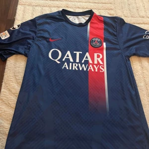 PSG Mbappé 7 matchtröja Nike blå - Paris Saint-Germain fotbollströja med Mbappé 7 på ryggen. Djupblå med röd och vit vertikal rand, PSG-emblem på bröstet och Qatar Airways-tryck. Tillverkad i lätt, ventilerande material. Snyggt mönster i tyget och klassisk Nike Swoosh i rött.