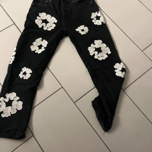 Levi's 501 svarta jeans denim tears - Svarta Levi's 501 jeans med vita blommiga broderier över hela byxorna. Klassisk rak passform och fem fickor. Jeansen är gjorda i slitstarkt denim och har en cool kontrast mellan svart tyg och vita detaljer. Perfekt för dig som vill sticka ut med en unik look.