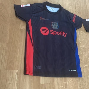 FC Barcelona Lamine Yamal tröja Nike - Snygg FC Barcelona matchtröja med Lamine Yamal och nummer 19 på ryggen. Tröjan är mörkblå med röda och blå detaljer, korta ärmar och tryck från Spotify och UNHCR. Tillverkad i lätt Dri-FIT material som andas, perfekt för fotboll eller häng med kompisar.