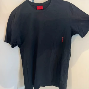 Svart t-shirt från Hugo Boss - Svart t-shirt från Hugo Boss med röd HUGO-logga tryckt på bröstet. Klassisk rund hals och korta ärmar. Tillverkad i mjuk bomull som känns skön mot huden. Snygg och enkel design som funkar till jeans eller shorts.
