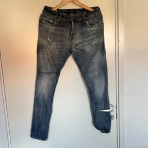 Blå slitna jeans från Jack & Jones - Säljer ett par blå jeans från Jack & Jones med snygga slitningar och distressed detaljer. Modellen har klassisk femficksdesign, normal passform och raka ben. Jeansen är tillverkade i jeansmaterial med ljus tvätt och coola slitningar på låren och knäna.
