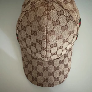 beige keps med GG-mönster - Snygg beige keps med klassiskt GG-mönster i brunt över hela kepsen. På sidan och baktill finns gröna och röda ränder som ger extra lyxig känsla. Justerbar baktill med tygrem. Materialet är bomull och polyester.