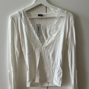 Vit långärmad blus med spets Gina Tricot - Supersöt vit blus från Gina Tricot med djup v-ringning och spetsdetaljer längs halsen. Långärmad och tillverkad i mjuk bomull som känns skön mot huden. Perfekt för dig som gillar romantiska vibes och vill ha en enkel men snygg topp i garderoben.