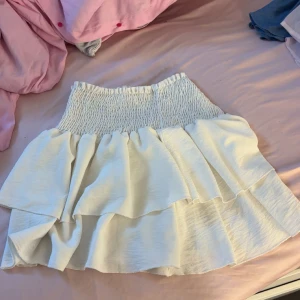 Vit volangkjol med smock - Supersöt vit kortkjol med bred smock i midjan och dubbla volanger. Kjolen är luftig och stretchig. Storlek M men passar även mig som bär xs-s🩵