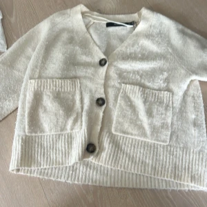 Beige kofta med knappar från Vera Moda - Mysig beige kofta från Vera Moda med v-ringning och tre stora knappar framtill. Koftan har två praktiska fickor och ribbade detaljer vid fickor och nederkant. Perfekt för lager-på-lager och chill dagar.