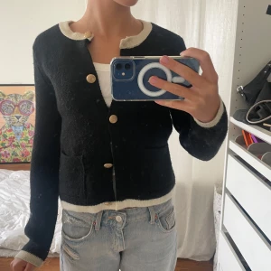 Svart kofta med guldfärgade knappar från Zara - Snygg svart kofta från Zara med kontrasterande vita kanter vid hals, ärmslut och nederkant. Koftan har guldfärgade dekorativa knappar och två fickor framtill. Stickad i mjukt material och har en klassisk, lite kortare passform som är lätt att styla med jeans.