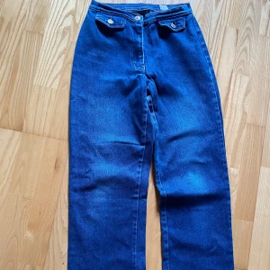 Blå raka jeans från Zoul, storlek 34 - Snygga blå jeans från Zoul i rak modell med hög midja. Klassisk femficksdesign med två fickor framtill med lock och knapp. Jeansen har en enkel, clean look. Perfekta för dig som gillar tidlös stil och vill ha ett par jeans som funkar till allt.