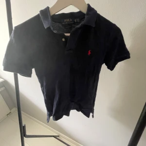Mörkblå Polo Ralph Lauren pike - Snygg mörkblå piké från Polo Ralph Lauren med klassisk krage och korta ärmar. Den har den ikoniska röda broderade loggan på bröstet och två knappar vid halsen. Passformen är normal och materialet är mjuk bomull. 140/152