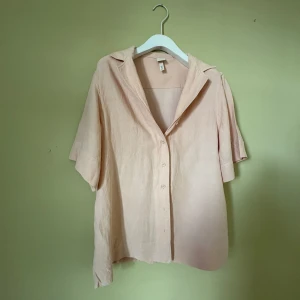 Beige linneskjorta med kort ärm - Snygg och luftig beige skjorta från H&M i linne med korta ärmar och klassisk krage. Skjortan har knappar framtill och en avslappnad passform som gör den perfekt för varma dagar. Enkel och stilren design som är lätt att matcha.