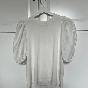 Vit puffärm topp från Kappahl XS - Supersöt vit topp från Kappahl i storlek XS med puffiga korta ärmar som ger en trendig och cool siluett. Toppen är gjord i mjuk bomull med glansiga ärmar som sticker ut och gör hela looken extra stylish. Perfekt att styla med jeans eller kjol för en chic vibe.