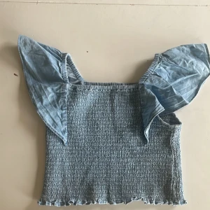 Ljusblå smockad topp från Kappahl - Supersöt ljusblå topp från Kappahl med smockad kropp och volangiga axelband. Toppen har en croppad passform och är tillverkad i mjuk bomull. Perfekt för dig som gillar romantiska detaljer och vill ha något som sticker ut med sina breda volangärmar.