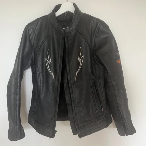 Svart skinnjacka Bullfighter - Säljer en svart skinnjacka från Bullfighter med broderade detaljer och logga på både ärm och rygg. Jackan har dragkedja framtill, ståkrage och coola vita broderier på bröstet. Perfekt för dig som vill sticka ut med en edgy look.