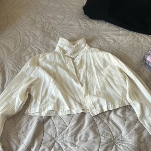 Randig croppad skjorta från H&M Divided - Snygg croppad skjorta från H&M Divided i ljusbeige och vit med diskreta ränder. Skjortan har lång ärm, klassisk krage och knappar framtill. 