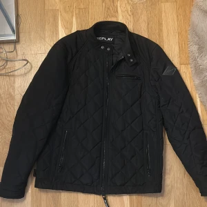 Svart quiltad jacka från Replay - Snygg svart quiltad jacka från Replay med ståkrage och dragkedja framtill. Jackan har flera fickor med dragkedja och ett stilrent Replay-märke på ärmen. Perfekt för dig som gillar en clean look med lite edge. Passar dig som vill ha en jacka med coola detaljer.
