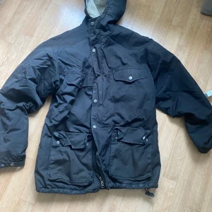 Svart vinterjacka från Fjällräven M - Säljer en svart Greenland Winter Jacket från Fjällräven i storlek M. Jackan har huva, flera stora fickor med knappar och dragkedja framtill. Insidan är fodrad med mjuk teddy och yttertyget är slitstarkt. Perfekt för kalla dagar och har en klassisk, rymlig passform.