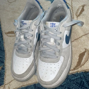 Nike Air Force 1 Athletic Club sneakers - Nike Air Force 1 Athletic Club sneakers i vitt och grått med blå detaljer och klassisk swoosh på sidan. Skorna har snörning, rund tå och platt sula. Ovandelen är i mocka och skinn, med broderad Athletic Club-logga på hälen. Perfekt för dig som gillar retrostil och streetwear.