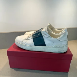 Valentino Garavani vita sneakers - Valentino Garavani sneakers i vitt skinn med marinblått band och nitar på hälen. Väldigt slitna och därför säljs för lågt pris.